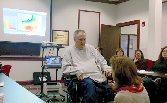 A man in a power wheelchair in a conference room talking with a woman kneeling before him. Behind him is a projected image of a pressure map showing a red hot spot. There are clinicians observing seated on the photo's perimeter.