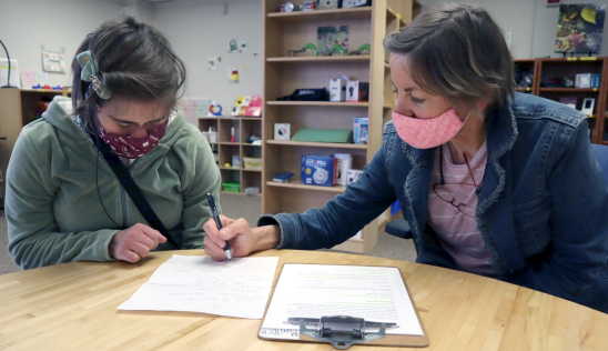 Two women work together at a table wearing masks, one assisting the other, eyes cast on paperwork.