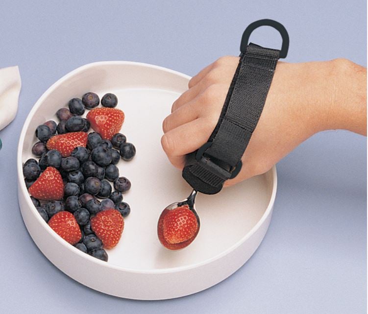 A hand wearing a velcro-style cuff that holds a spoon with a strawberry. There is a loop at the back of the hand. The hand is scooping from a wide-brimmed bowl containing blueberries and strawberries.