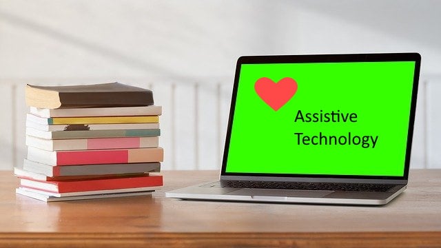 A stack of books on a table with an open laptop displaying a heart and the words "assistive technology"