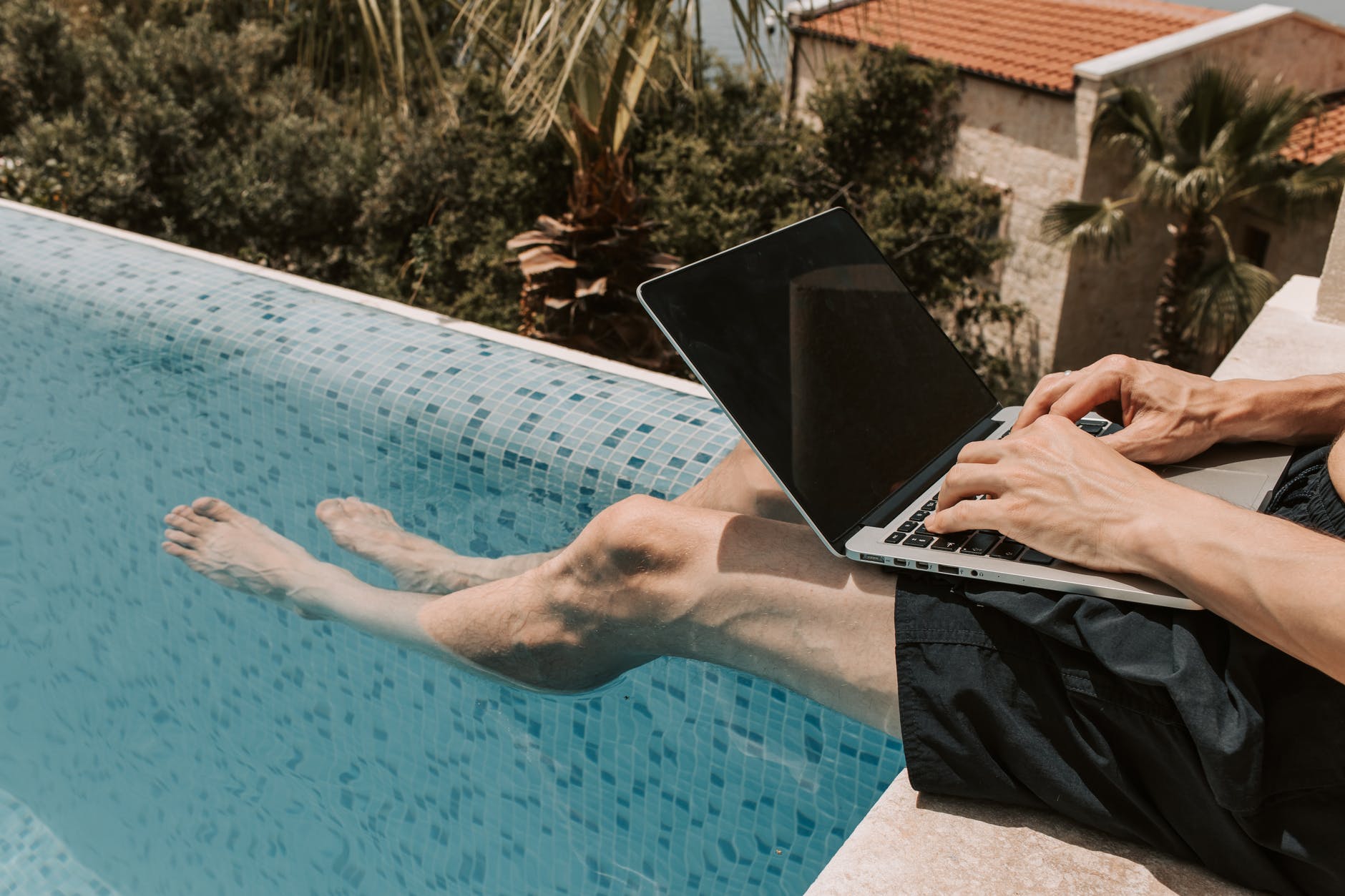 Crop photo of man dangling his legs into a swimming pool while using a laptop.