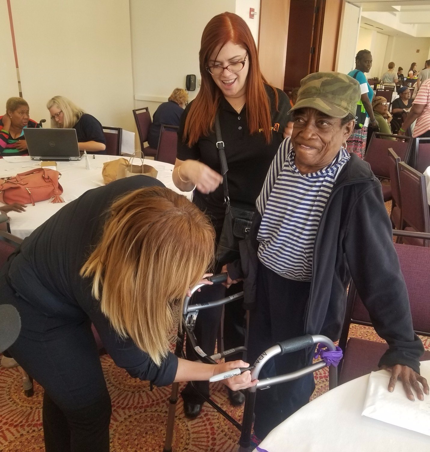 A woman Caribbean hurricane refugee with a walker receiving equipment assistance from two women in a conference center room.