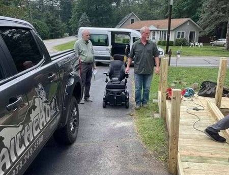 Two men with a power wheelchair in a driveway next to a ramp under construction.
