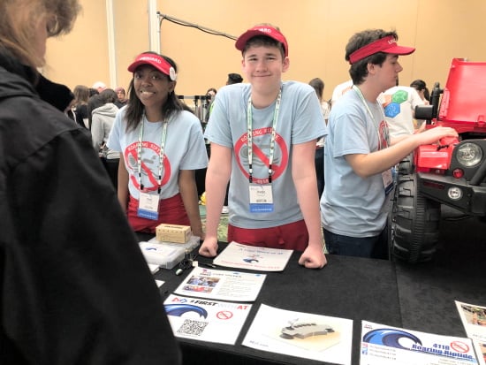 Two smiling members of the Roaring Riptide robotics team of Florida University, an African-American young woman and a Caucasian young man. An additional teammate stands to the side working on a large toy car. All are behind a display table in an expo hall.
