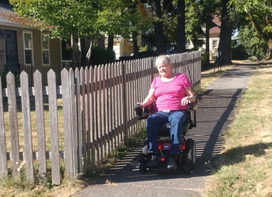 An older woman travels a sunny sidewalk along side a picket fence in her power wheelchair smiling.