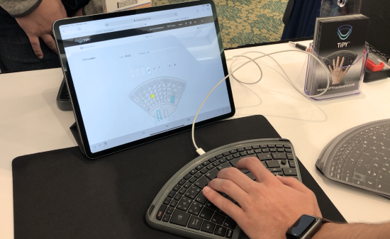 A sleek flat wedge-shaped keyboard with wrist support and keys that fan out above like concert seating. A man's hand is typing with the aid of a tablet and app that demonstrates which finger to use for specific letters and functions.