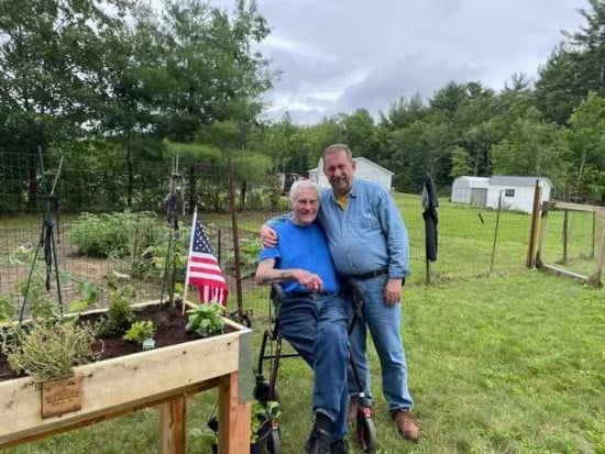 Two men in a backyard, one seated in a rollator, the other embracing him, are next to an accessible raised garden bed with an American flag planted.