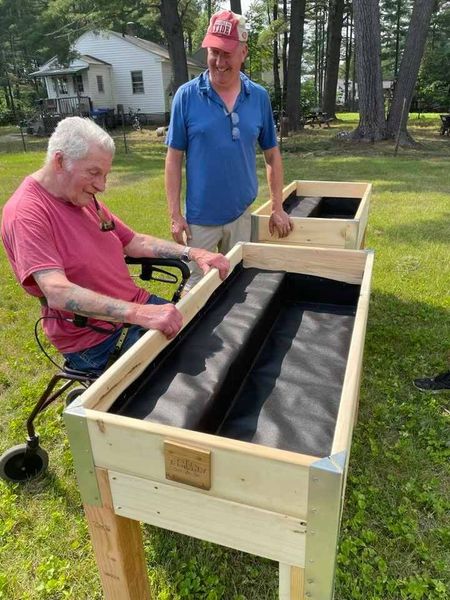 A man seated in a rollator examines brand new raised garden beds while another man smiles on.