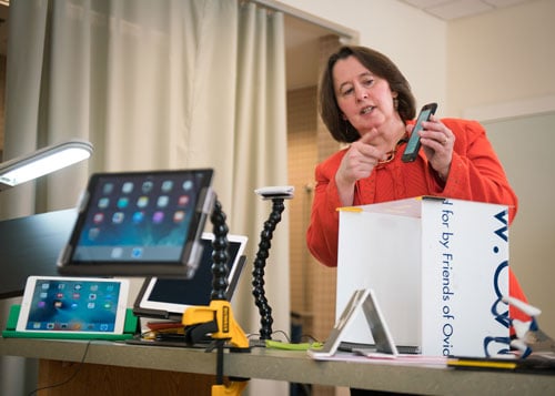 Una mujer detrás de una mesa en la que se exponen tabletas montadas de distintas formas, incluida Loc-Line con una pinza, sostiene y señala un teléfono inteligente.
