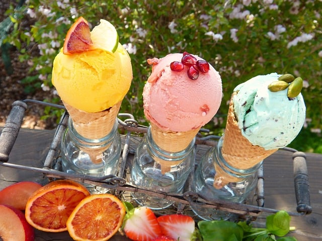 Three ice cream cones held upright in Ball Jars supported by an antique jar-holding tray. They are outside on a wooden table with cut fruit in front and flowers in the background.